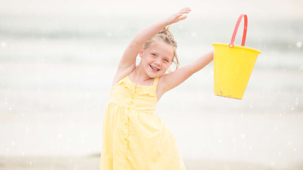 Ein kleines Mädchen in einem gelben Kleid mit einem gelben Eimer in der Hand am Strand Ein kleines Mädchen in einem gelben Kleid mit einem gelben Eimer in der Hand am Strand